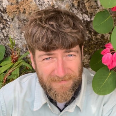 A white man in a white shirt against a background of rock and plants